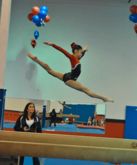 Alumni spotlight profile for Abigail Panilan-Yan. The section features a personal testimonial about her journey from a Level 10 gymnast to a coach and business owner. Two photos are included: on the left, a younger Abigail performs a split leap over a balance beam in a red and black leotard; on the right, a modern portrait shows her smiling and holding nail polish bottles for her business, 'Abigail Nail Lab'.