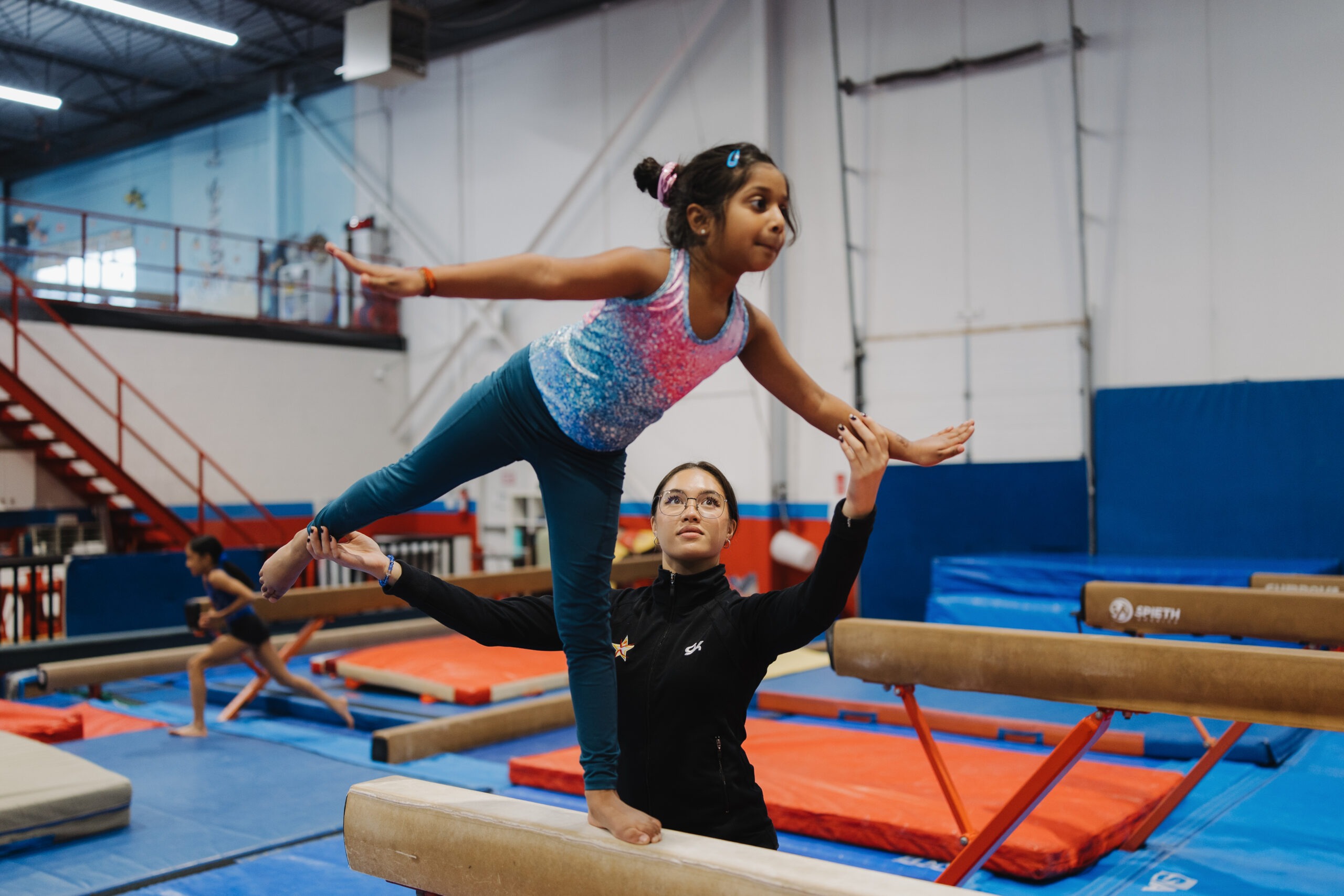 Young gymnast in pink-blue leotard balancing on beam with coach.