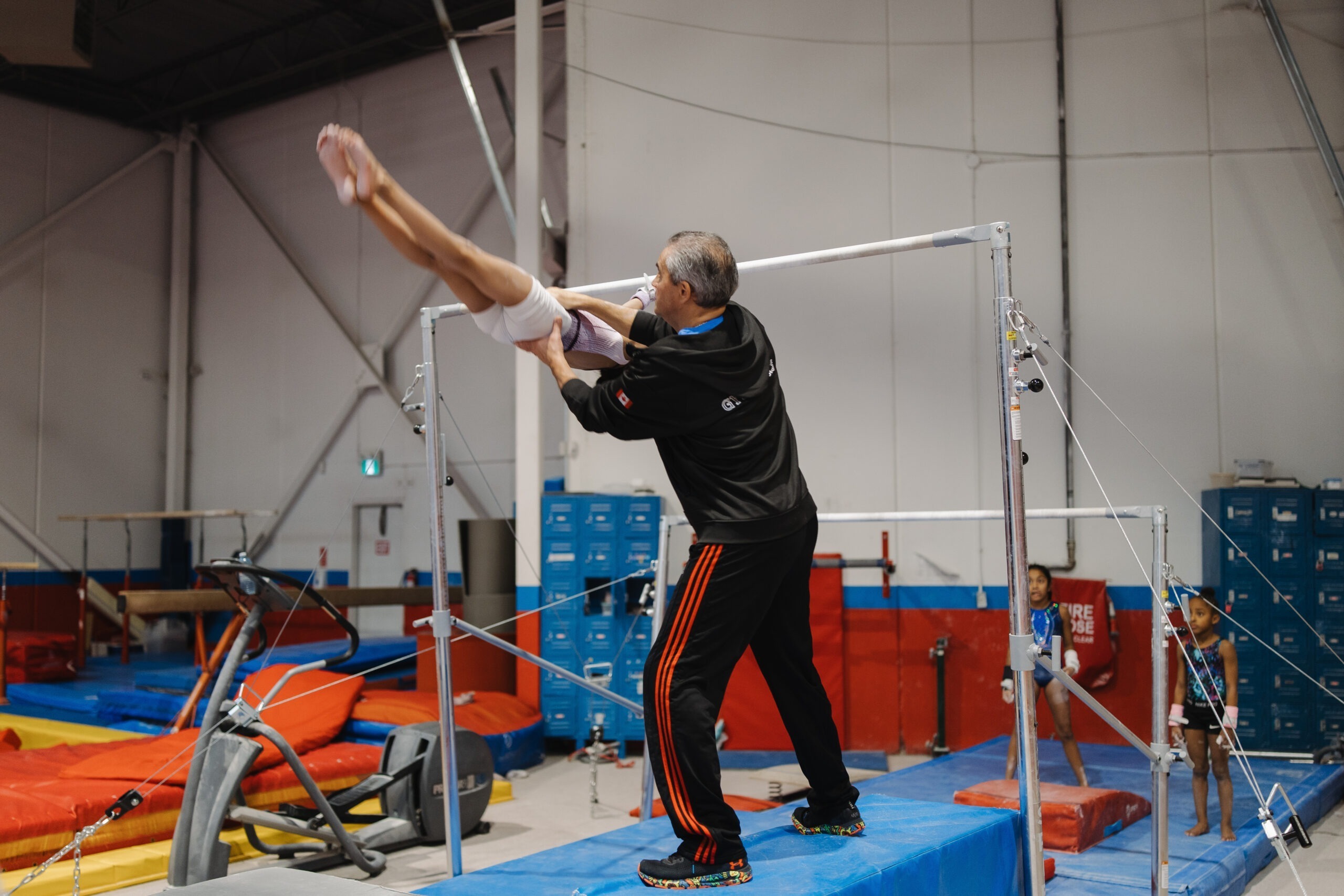 A senior gymnastics coach supporting a young athlete as they perform a horizontal release move on the uneven bars.