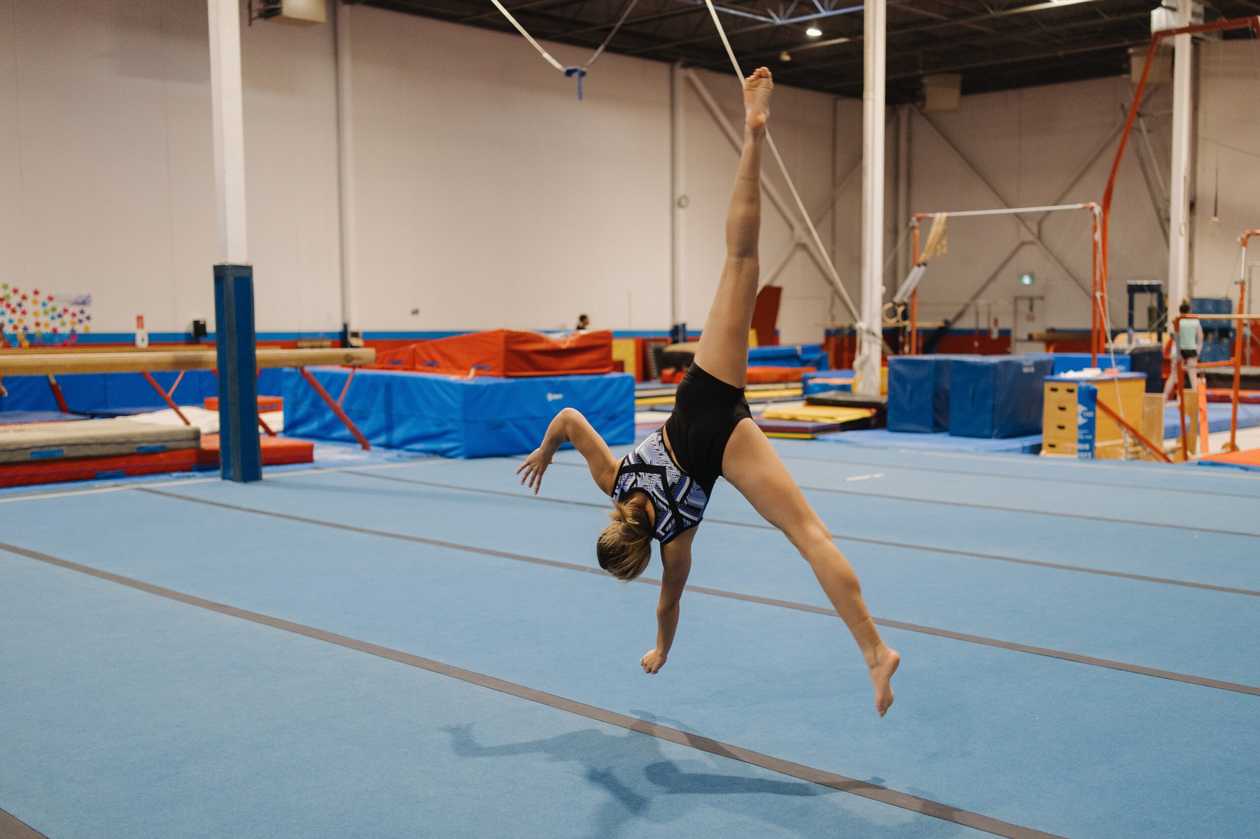 Teen gymnast in black-blue leotard doing one-handed cartwheel on mat.