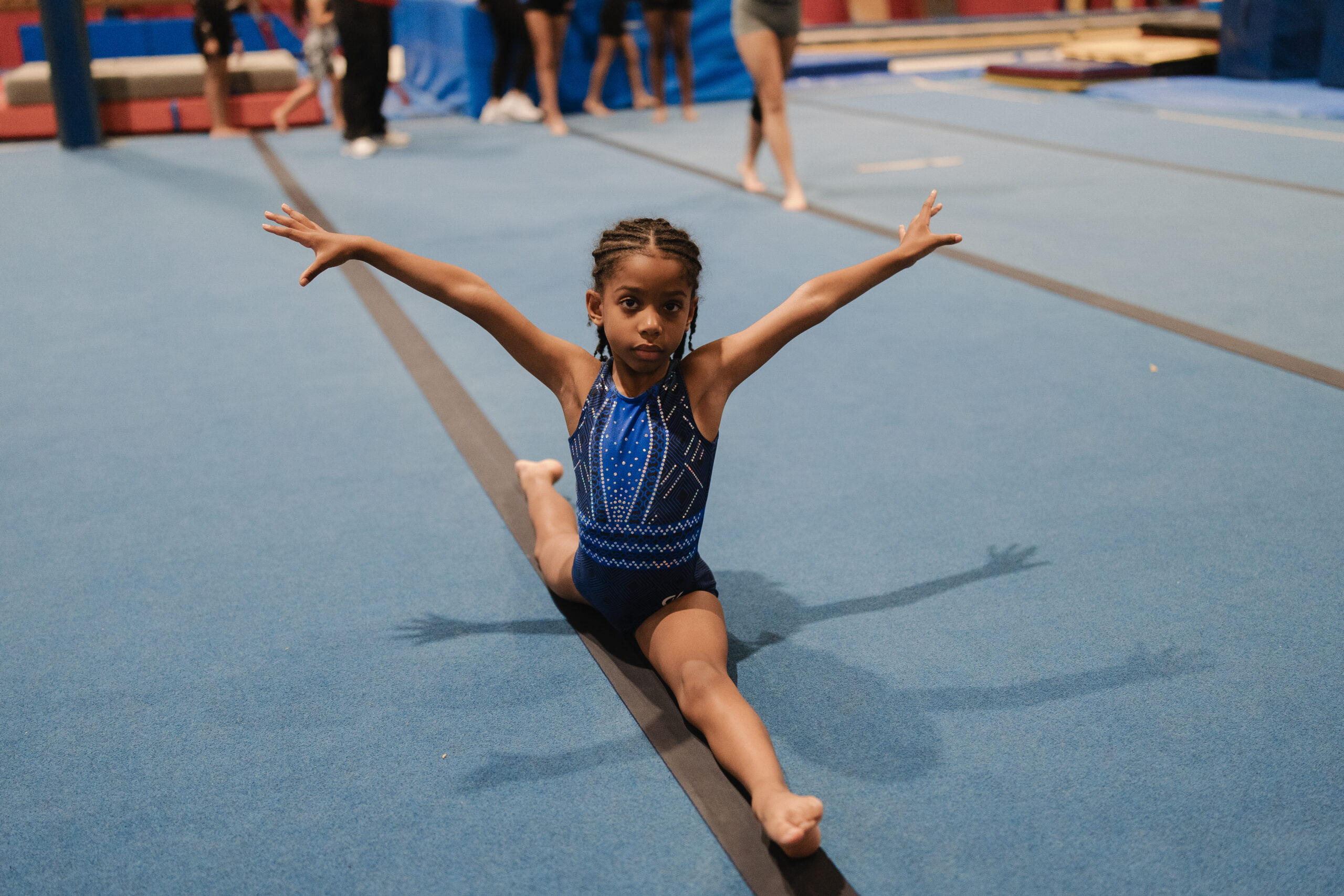 A young gymnast in a blue leotard performing a full middle split on a floor beam for the Gym Flex program card.
