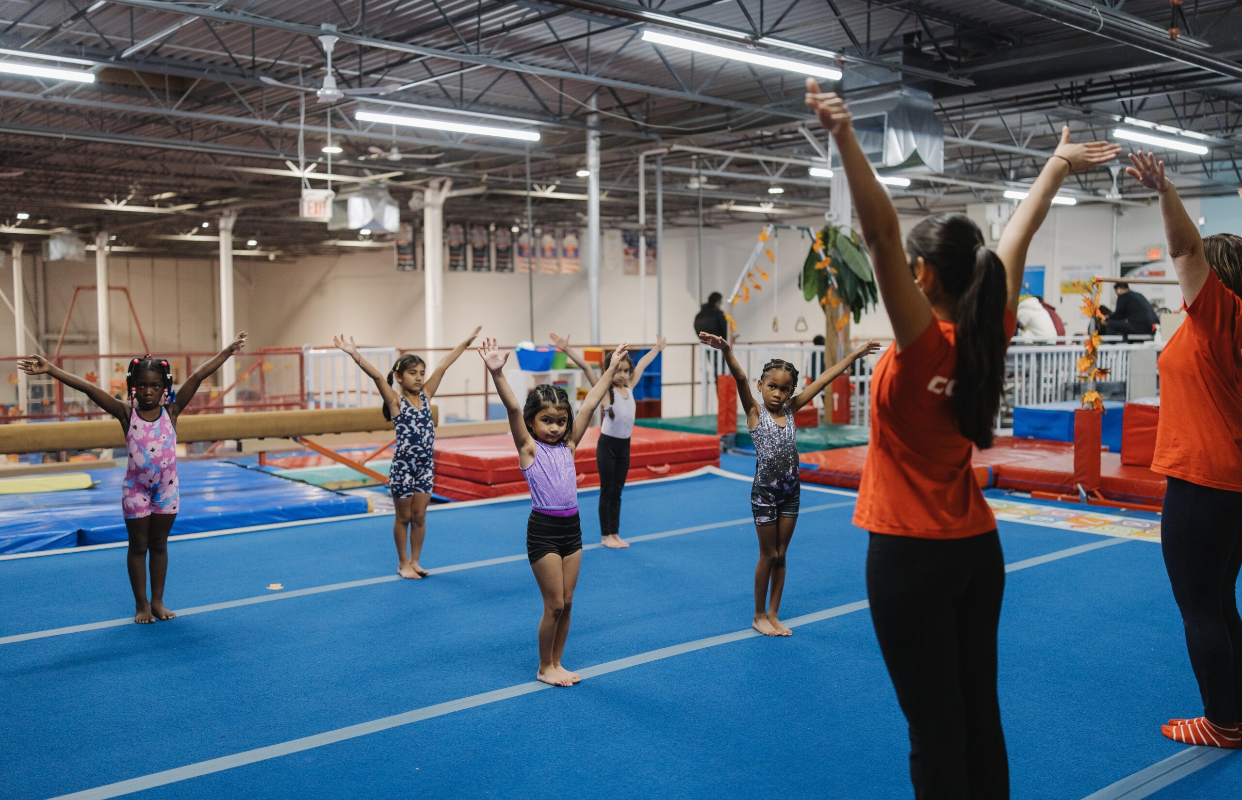 A group of young children in a gym standing on blue floor mats with their arms raised during a Tigers Kinder Camp session.