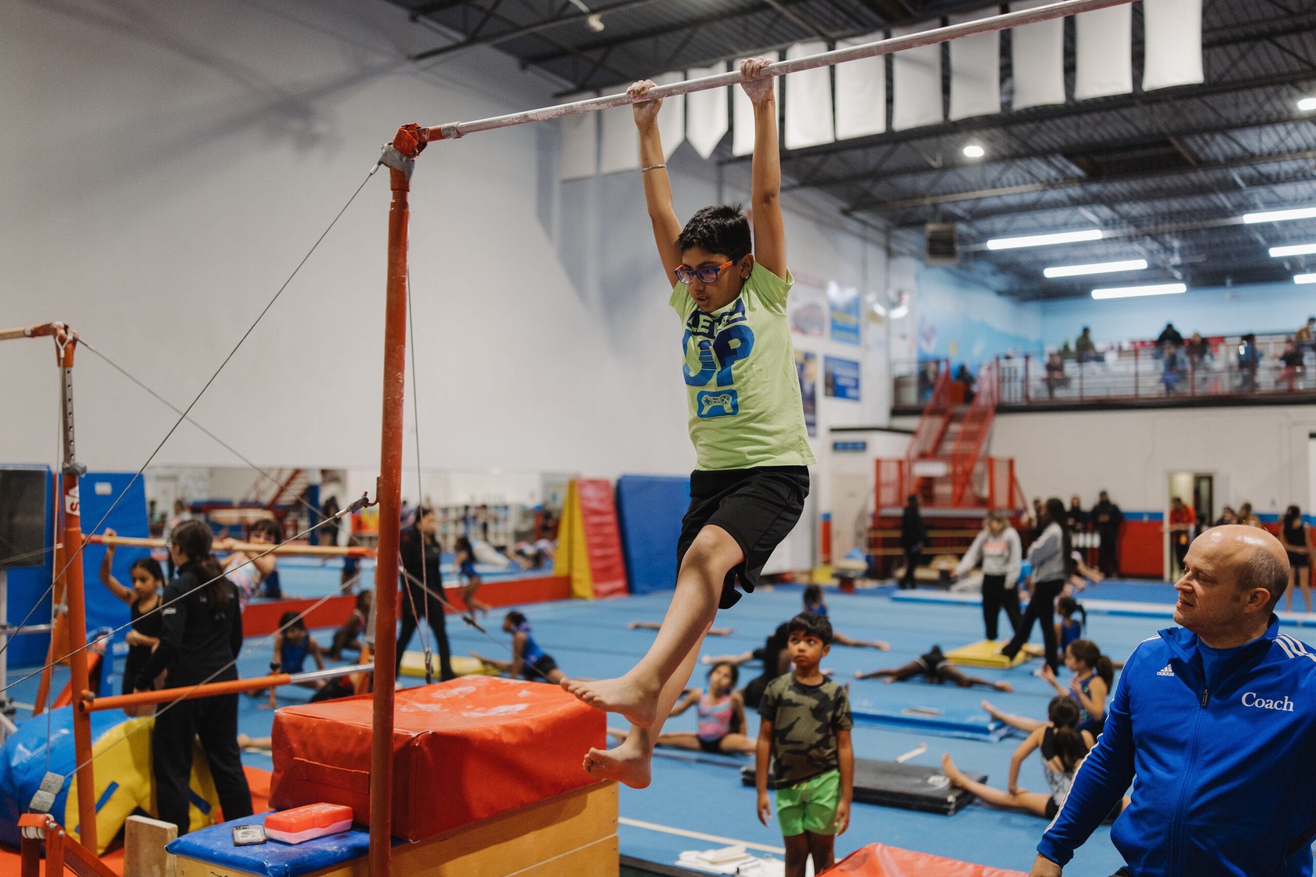A young boy in a green shirt practicing on a high bar at a gymnastics facility while a coach observes. Below the image is a description for 'Boys GymFit Intro | Levels 1 & 2' for ages 6–14, detailing a high-energy program focused on strength and coordination, with registration buttons for Junior and Senior levels.