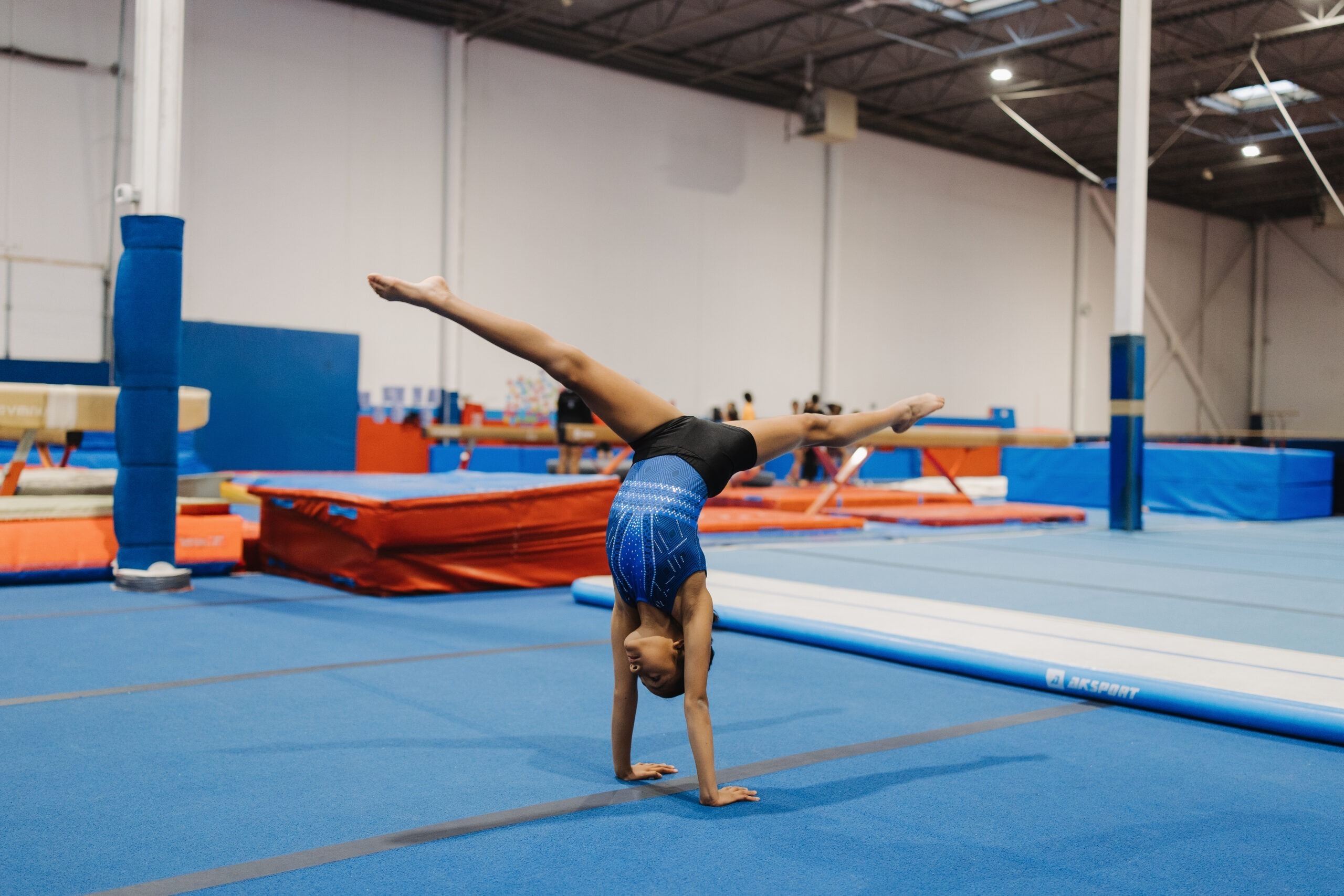 A young gymnast in a blue leotard performing a handstand with a leg split on a blue floor mat. Below the image, text describes the 'Intermediate Tumbling | Levels 5-8' program for ages 6 and up, focusing on mastering advanced skills like walkovers, roundoffs, and handsprings, with a red Register button at the bottom.