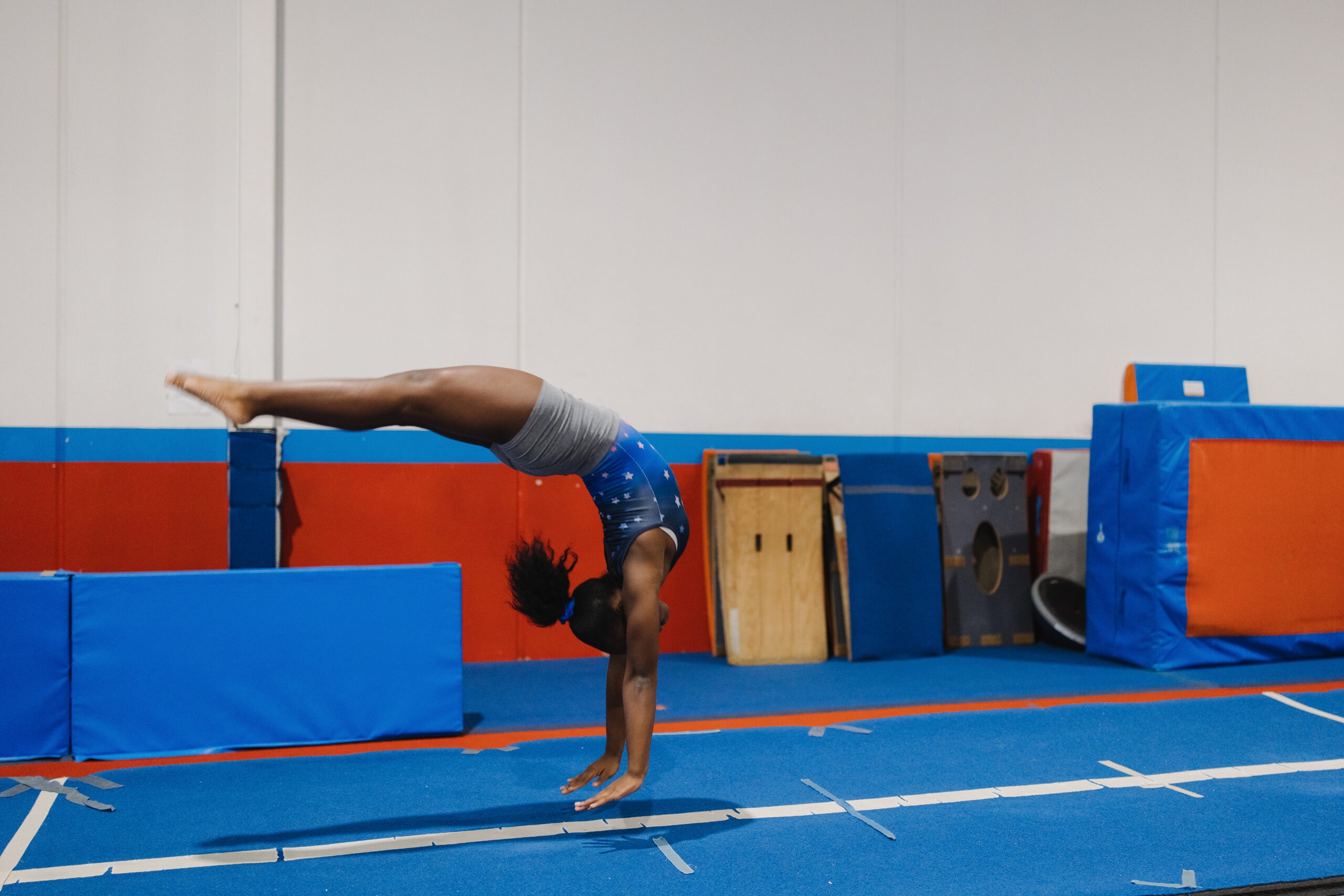 A young gymnast in a blue star-patterned leotard performing a back handspring on a blue floor mat. Below the image, text describes the 'Advanced Tumbling | Levels 9-10' program for ages 8 and up, focusing on high-level skills like back tucks, layout step-outs, and aerials, with a red Register button at the bottom.