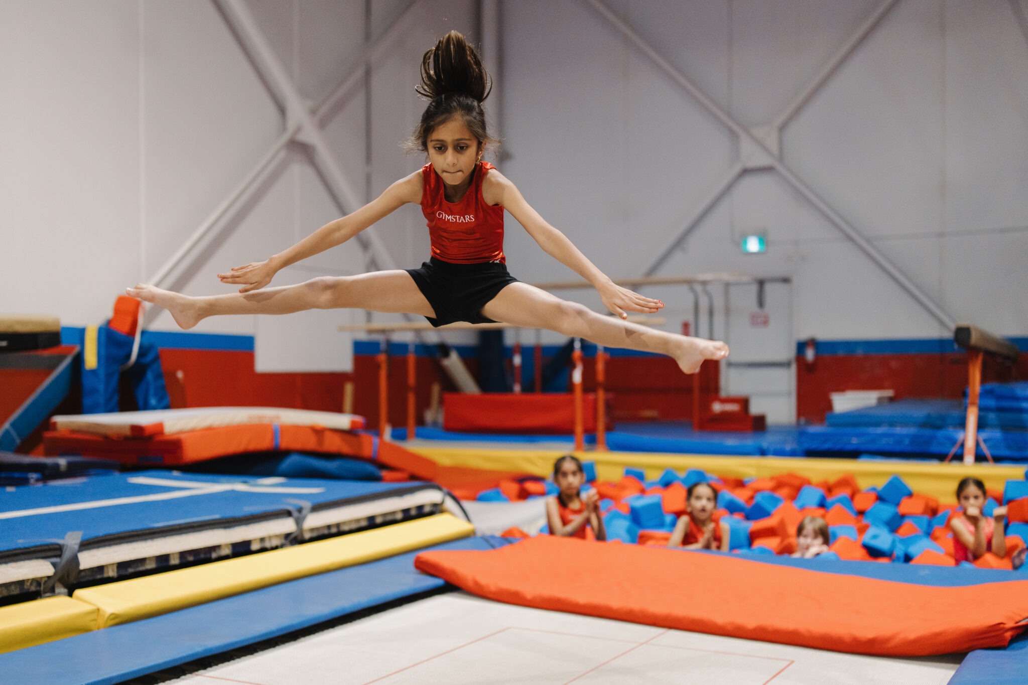 Gymnast in red-black leotard performing straddle jump above mat.
