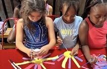 Three young campers sitting at a red table and focused on an arts and crafts project using paper and glue.