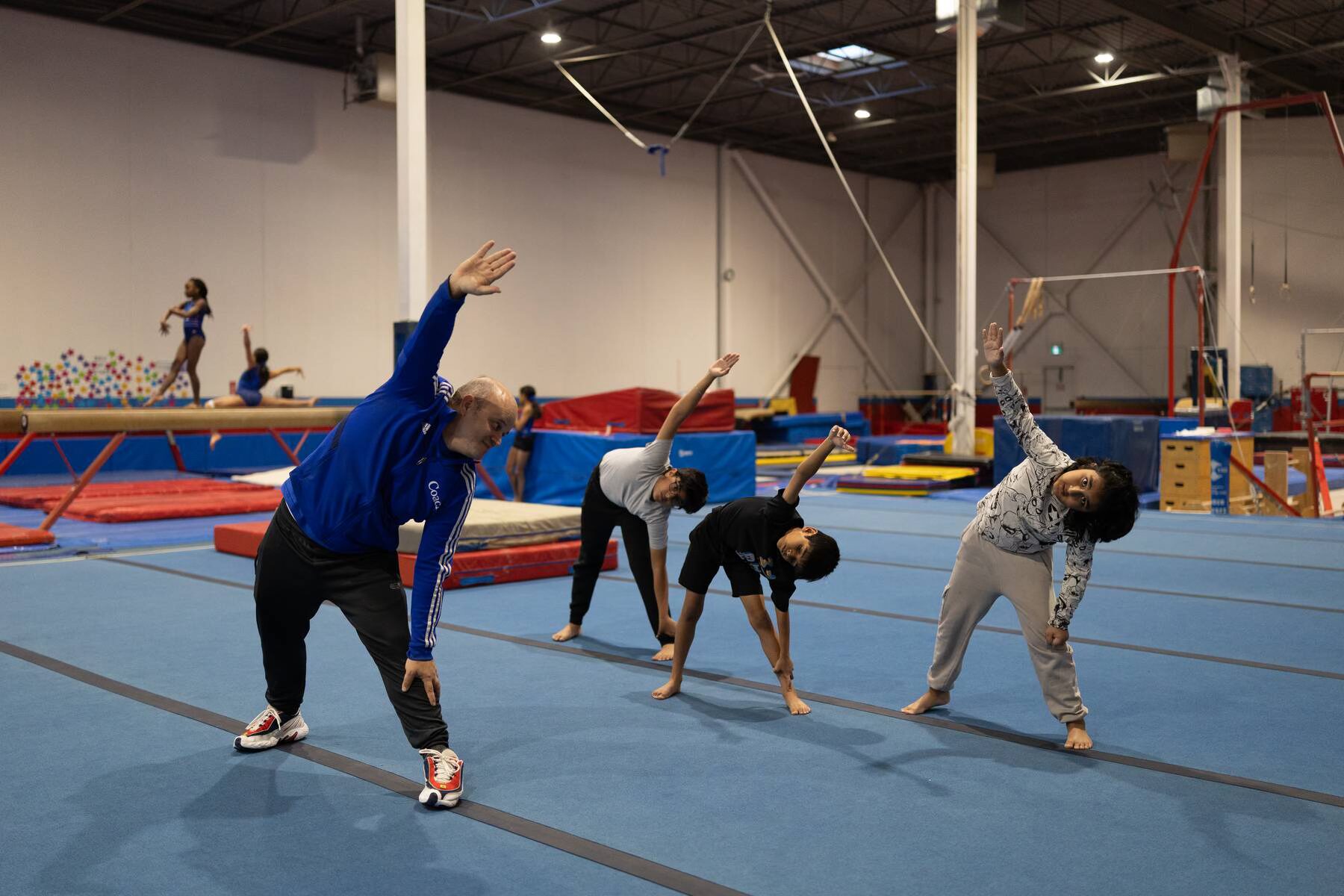 A gymnastics coach in a blue jacket leading three boys in side-stretch warm-ups on a blue floor mat. Below the image, text describes the 'Boys GymFit | Levels 3 & 4' program for ages 6–14, emphasizing core skills, strength work, and discipline, with registration buttons at the bottom.