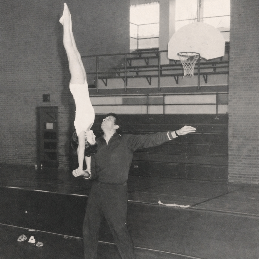 A vintage black-and-white photograph of a young Paula Lockwood performing a steady handstand while being balanced by a coach’s hand in a gymnasium.