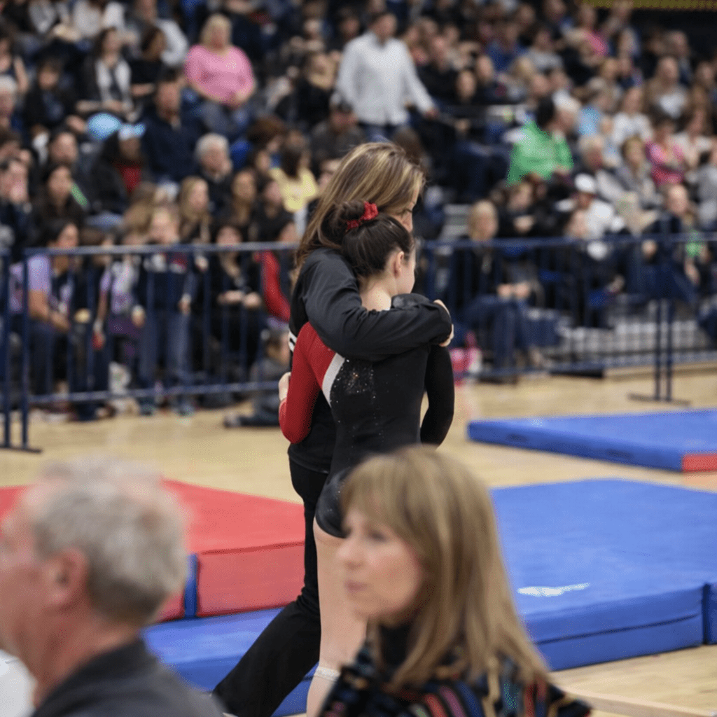 A group of young female gymnasts in blue and black leotards sitting together on a gym floor and smiling, representing the focus on building confidence and lasting friendships.