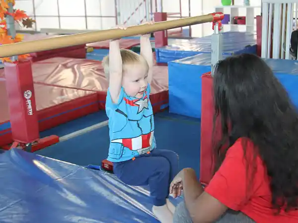Baby in Captain America shirt hanging from a low bar, supervised on a blue mat.