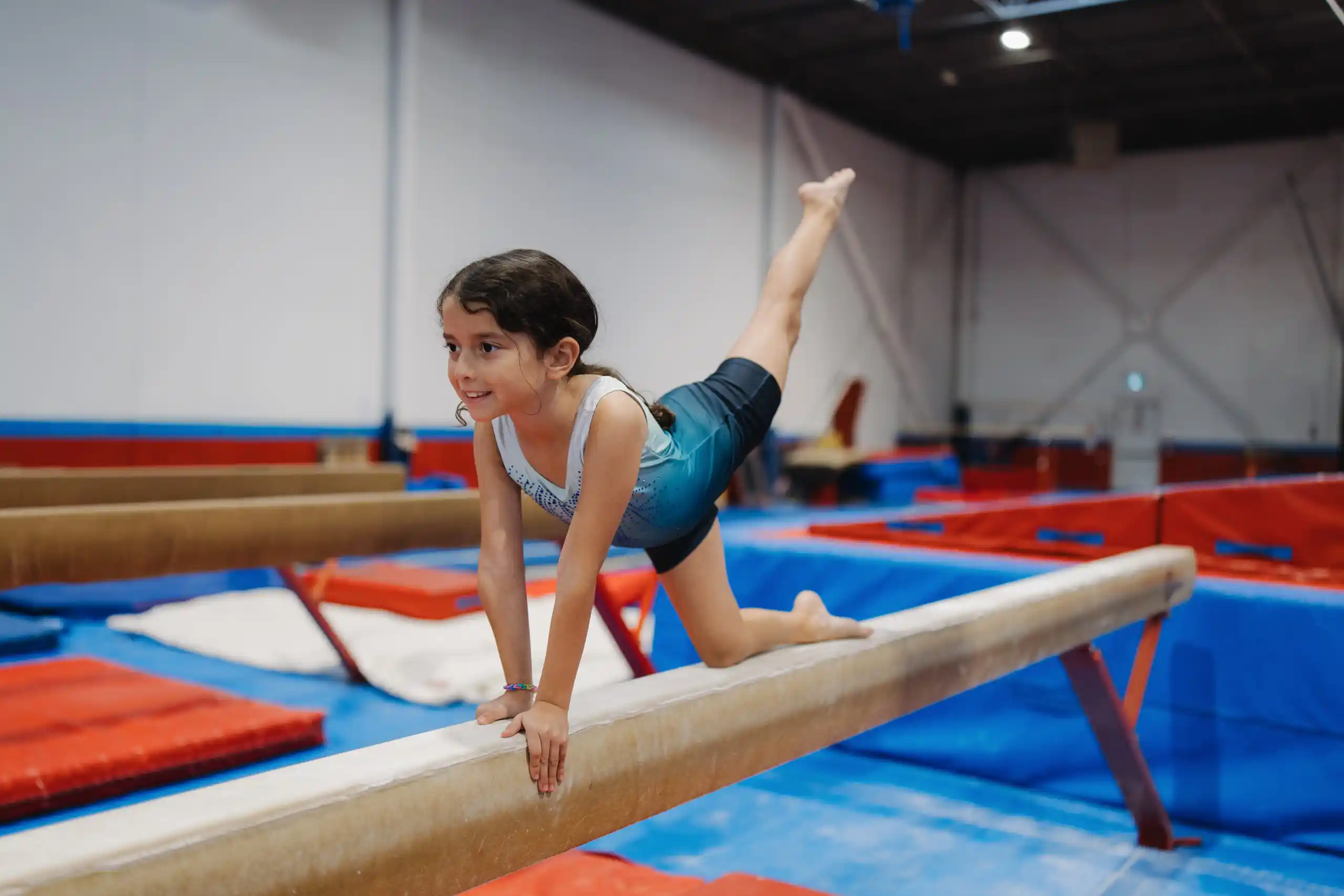 A young girl in a blue leotard practicing a balance scale move on a gymnastics beam in a gym.
