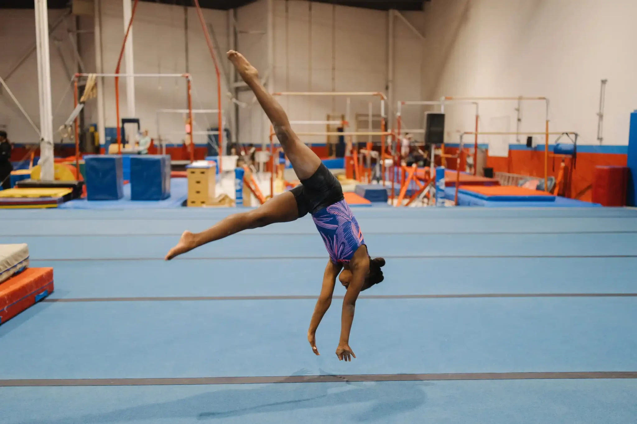 A young gymnast performing a cartwheel or tumbling pass on a long blue floor mat in a large gym.