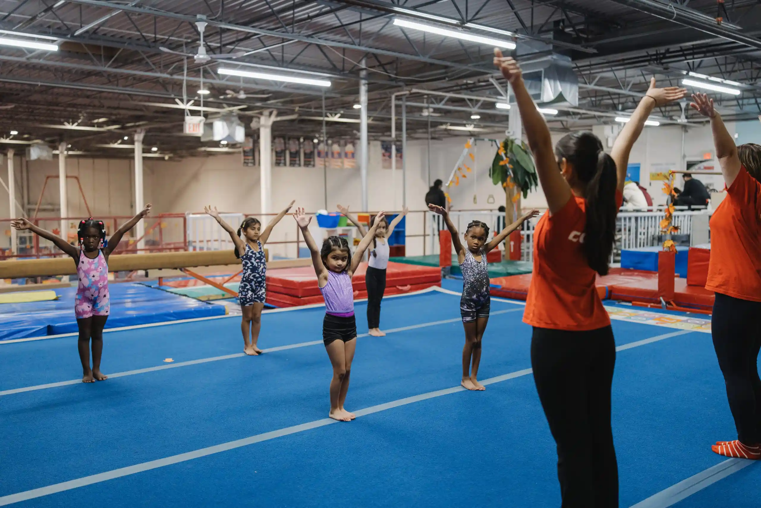 A group of young children in leotards standing on a blue gymnastics mat with their arms raised in a "ta-da" pose, led by a coach.