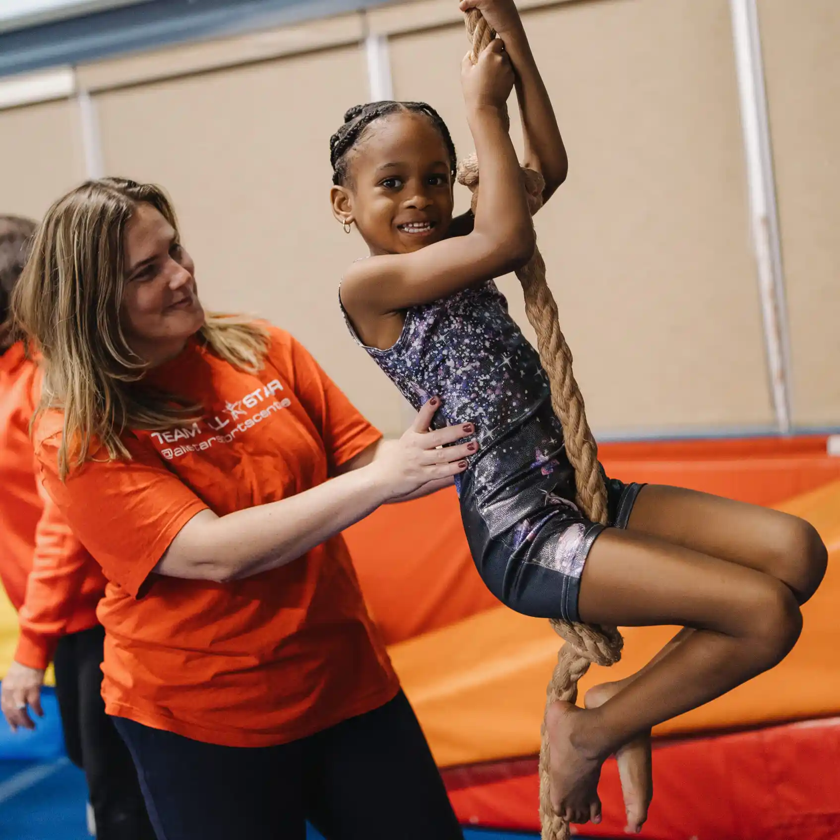 Girl in patterned leotard climbing rope, supported by coach in orange t-shirt.