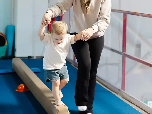 Toddler in white and blue walking a low balance beam, holding a parent's hand.