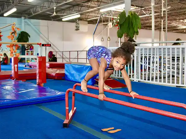 Girl in blue-purple leotard squatting on red parallel bars.