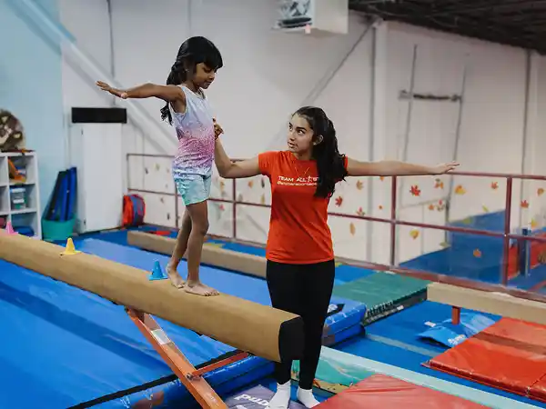 Girl in colorful leotard balances on beam, spotted by coach in orange shirt.