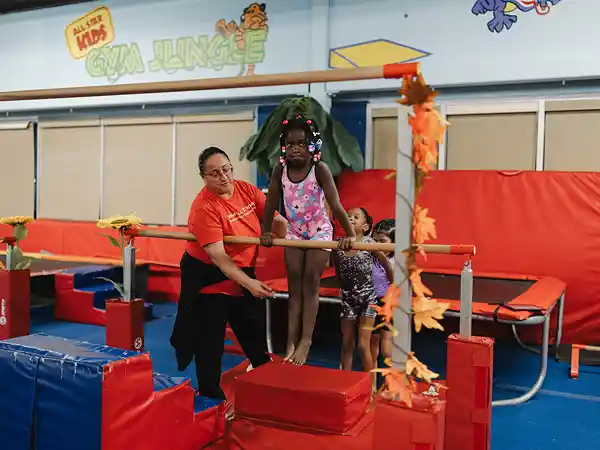 Girl in floral leotard on red block, holding bar, supervised by coach.
