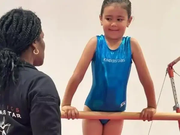 Girl in blue "Kinderstars" leotard resting on bar, looking at coach.