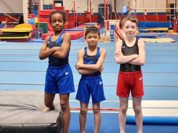 Three young male gymnasts standing side-by-side with their arms crossed in a professional gymnastics facility, representing the introductory competitive pathway that focuses on increased strength training and attention to detail.