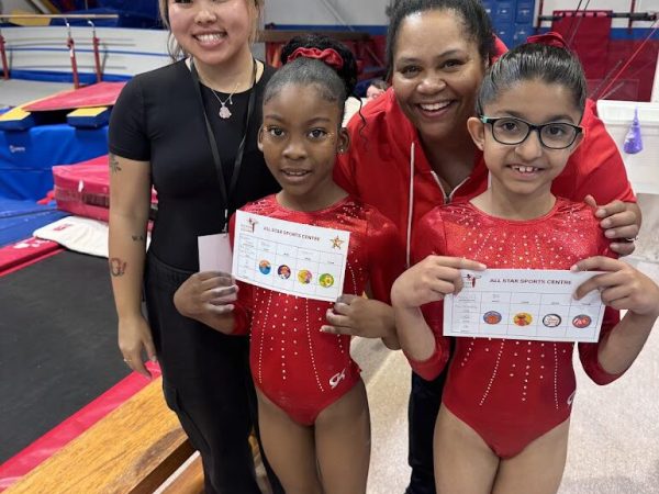 A female gymnast in a red leotard performing a high-altitude vault over a springboard and table in a professional competition arena. This advanced program focuses on refined technique and precision as athletes compete in JO Levels 3–9, requiring 12 to 20 hours of training per week and participation in three qualifying meets plus championships.