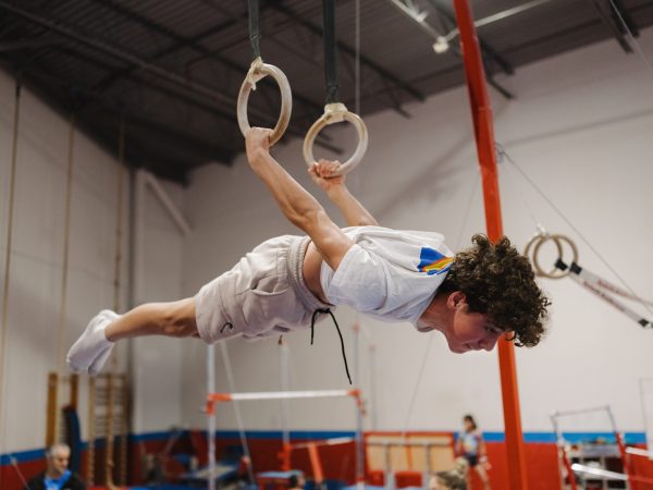 A young male gymnast performing a horizontal planche hold on the still rings in a professional gymnastics facility, illustrating the advanced skills and strength training required for regional competitions.