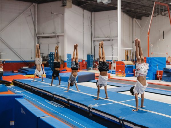 A young male gymnast in a red, white, and blue singlet performing a focused handstand on the floor mats of a professional gymnastics facility. This image represents the foundational training and discipline required for the Men's Artistic Gymnastics (MAG) program.