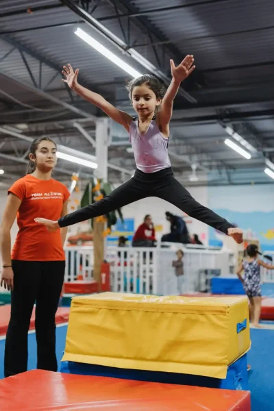 A young girl performs a straddle jump off a foam block while a coach supervises.
