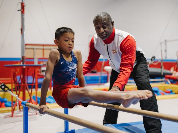 A young male gymnast in a red and blue singlet performing an L-sit hold on the parallel bars while being guided by a coach.